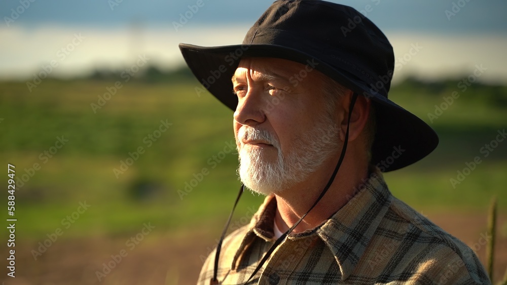 Close up portrait of senior caucasian good looking wise man farmer in a hat looking at the side, turning face to the camera in a field.