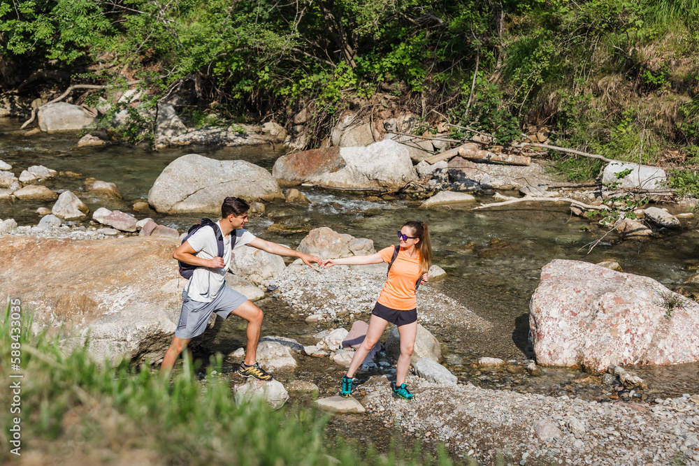 Active young hikers, man and woman walking over a mountain stream flowing among large beautiful rocks. Concept of nature exploring.