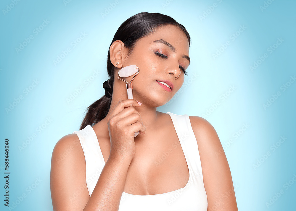 Asian Relaxed young woman standing smiling with closed eyes while using rose quartz face roller. Isolated on the blue background. Asian Relaxed young woman standing smiling with closed eyes while using rose quartz face roller. Isolated on the blue background.
