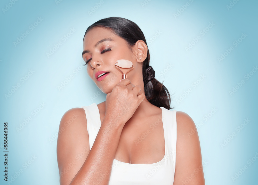 Asian Relaxed young woman standing smiling with closed eyes while using rose quartz face roller. Isolated on the blue background. Asian Relaxed young woman standing smiling with closed eyes while using rose quartz face roller. Isolated on the blue background.