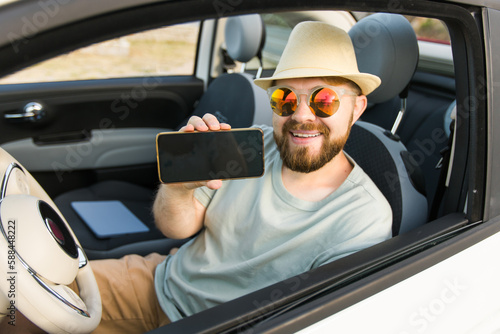 Smiling bearded man in sunglasses showing mobile phone with empty screen for mock up sitting on driver seat in new luxury car cabriolet, holding gadget in hand, selective focus on device
