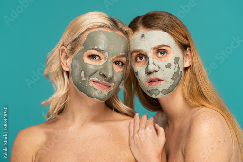 redhead and blonde women with clay mask on faces looking at camera isolated on turquoise.