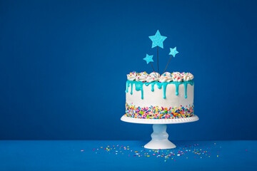 White birthday drip cake with teal ganache, star toppers over dark blue background