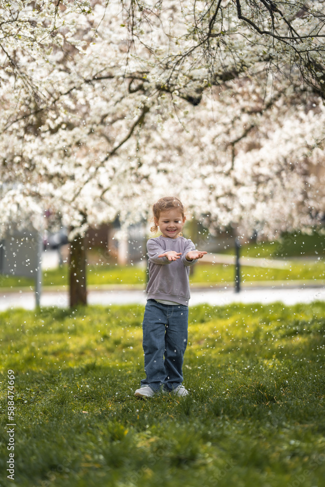 Little girl stands under a blooming apple tree. The wind blows and flower petals fly like snow in Prague park, Europe Little girl stands under a blooming apple tree. The wind blows and flower petals fly like snow in Prague park, Europe