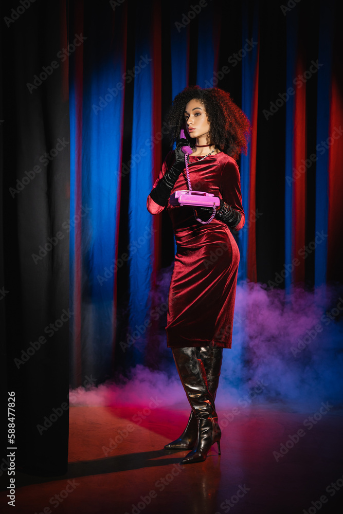 Full length of elegant african american model talking on telephone on blue background with light and smoke. Full length of elegant african american model talking on telephone on blue background with light and smoke.