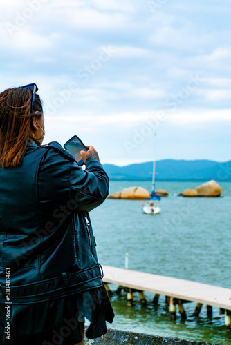 Mulher turista tirando foto de praia com veleiro e pedras ao fundo Coqueiros, Florianópolis, Santa Catarina, Brasil photo