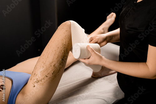 Bandage wrapping process after applying scrub on legs of girl for styx wrapping and banding anti-cellulite procedure in spa. Masseur is doing scrub massage for skin cleaning in spa.