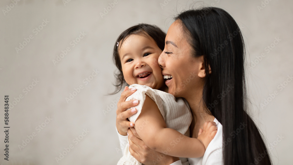 Joyful Asian Mom Hugging Baby Daughter Over Gray Wall Indoor Joyful Asian Mom Hugging Baby Daughter Over Gray Wall Indoor