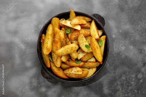 Roasted potatoes with herbs and spices. Baked potato wedges in frying pan on dark stone background. Top view with copy space.