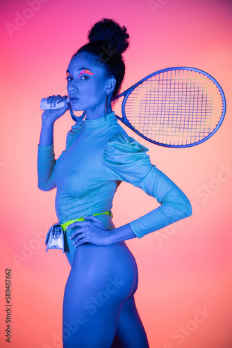 Trendy african american woman in bodysuit holding tennis racket on pink background with neon light. © LIGHTFIELD STUDIOS