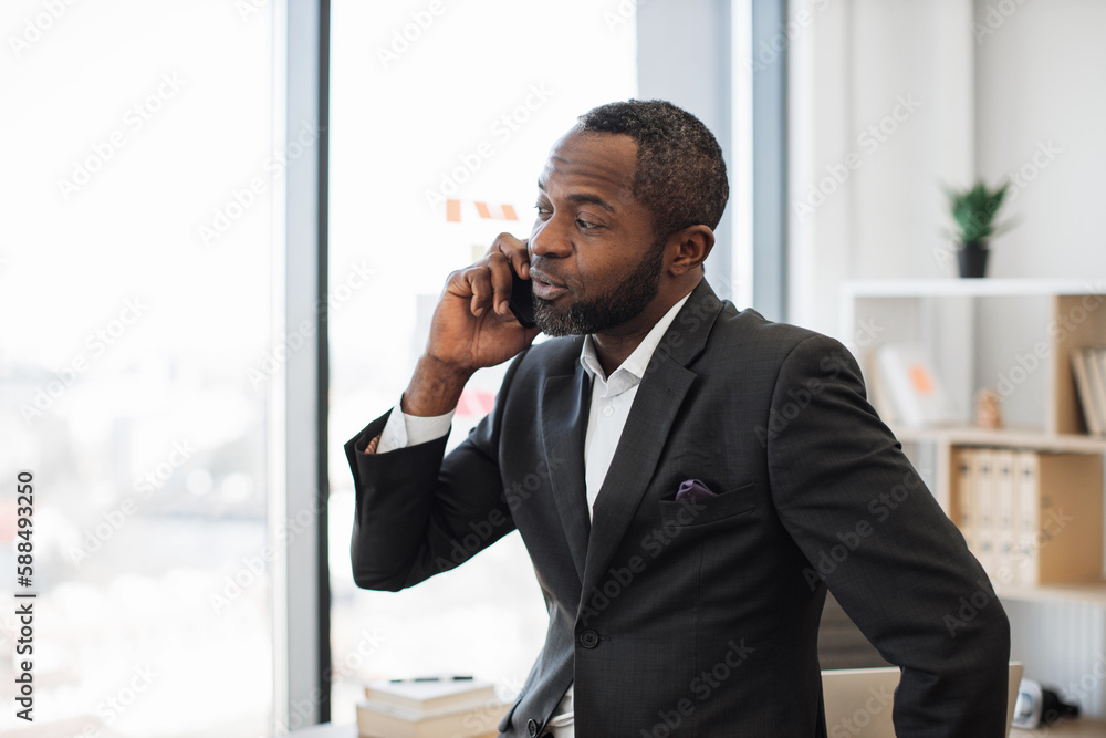 Fototapeta premium Stylish multiracial man in business suit talking on modern smartphone while staying indoors against office background. Chief executive having conversation with partner while waiting in corner office.