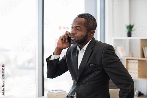 Stylish multiracial man in business suit talking on modern smartphone while staying indoors against office background. Chief executive having conversation with partner while waiting in corner office.