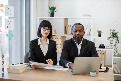 Multicultural white-collar male worker with laptop and his female caucasian colleague with paper sheets posing together at office table in meeting room. Confident partners planning corporate event.