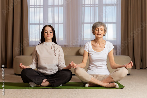A grandmother and her granddaughter are practicing yoga in a modern beige studio. An elderly woman in excellent physical shape is teaching and sharing her experience in yoga with the young girl.
