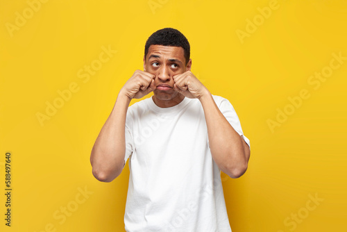 young sad guy african american in white t-shirt is crying on yellow isolated background, unhappy man is depressed