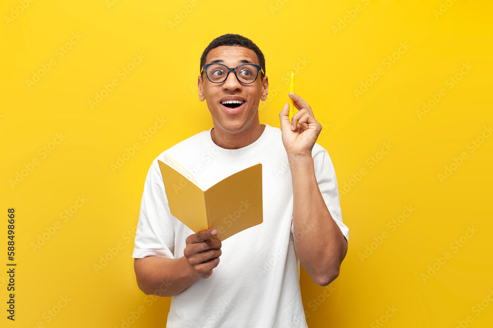 pensive guy african american student in white t-shirt and glasses writes in notebook and plans on yellow background pensive guy african american student in white t-shirt and glasses writes in notebook and plans on yellow background