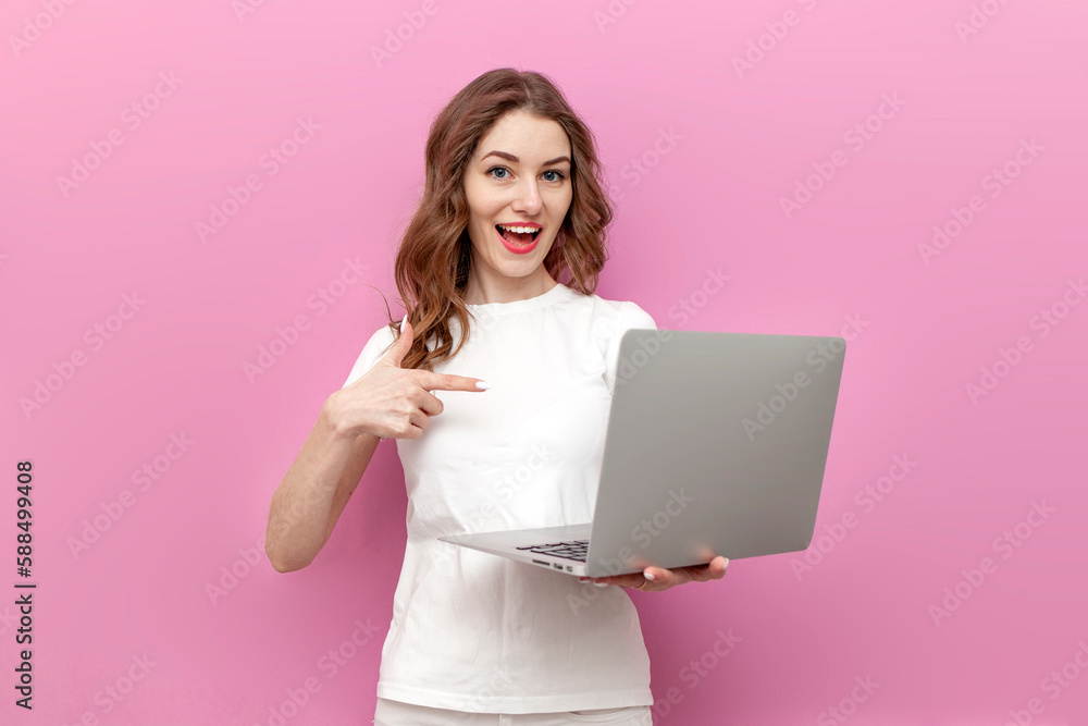 young cute woman in white t-shirt points finger at laptop and rejoices on pink isolated background young cute woman in white t-shirt points finger at laptop and rejoices on pink isolated background