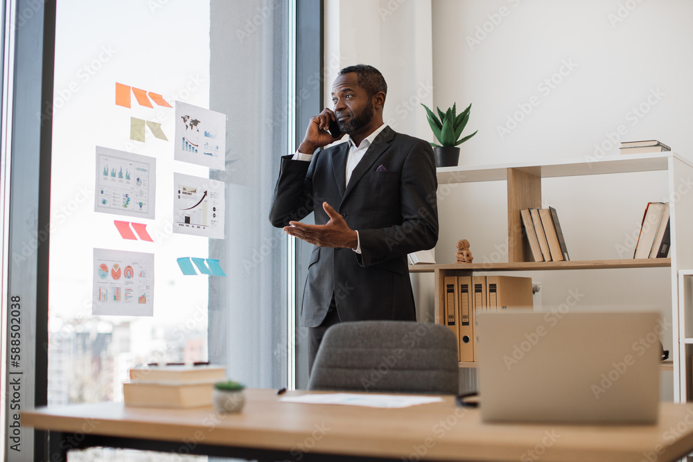 Multiethnic businessperson in formal outfit having conversation on cell phone while standing out against office background. Confident male employer thinking over speaker's words in cozy workplace. Multiethnic businessperson in formal outfit having conversation on cell phone while standing out against office background. Confident male employer thinking over speaker's words in cozy workplace.