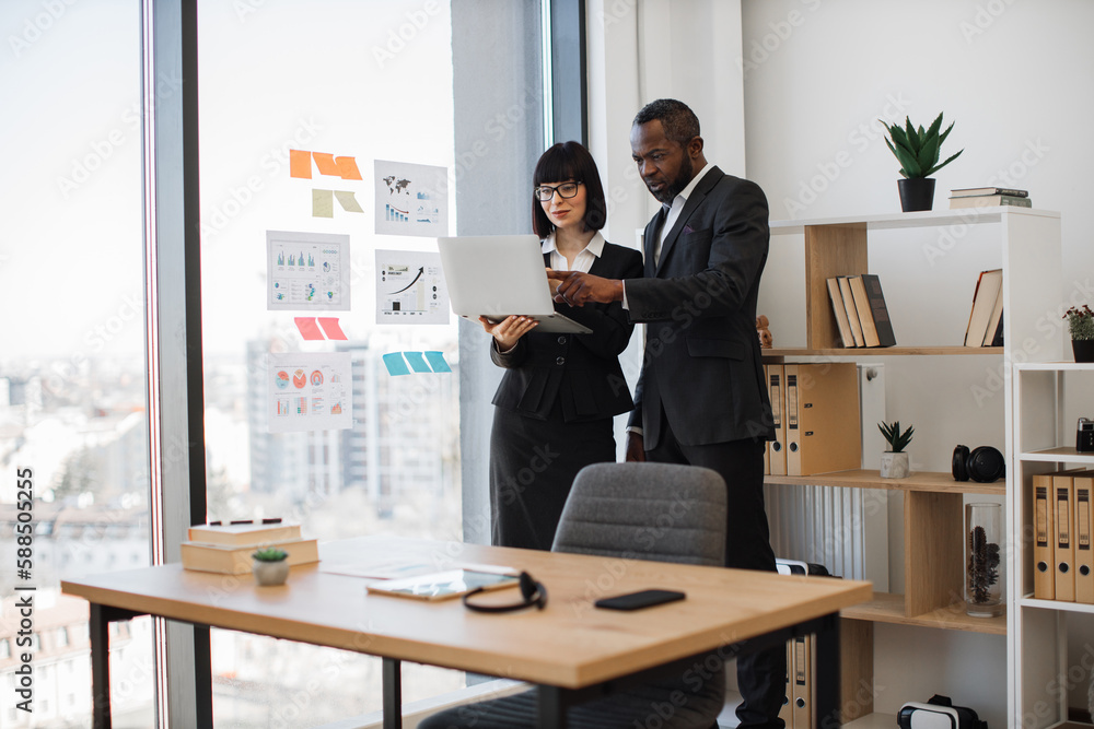 African american male in formal wear pointing at computer screen held by caucasian female in spectacles standing behind office table. Business partners working together on research plan indoors. African american male in formal wear pointing at computer screen held by caucasian female in spectacles standing behind office table. Business partners working together on research plan indoors.