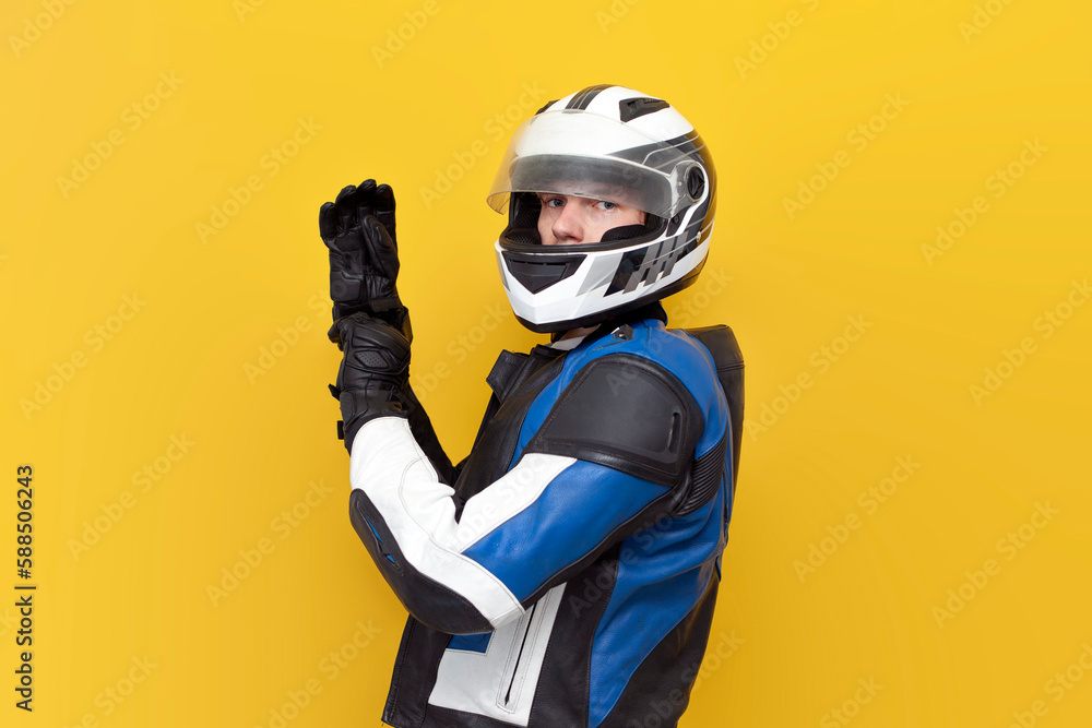 young guy motorcyclist in leather jacket and helmet puts on protective gloves on yellow isolated background young guy motorcyclist in leather jacket and helmet puts on protective gloves on yellow isolated background