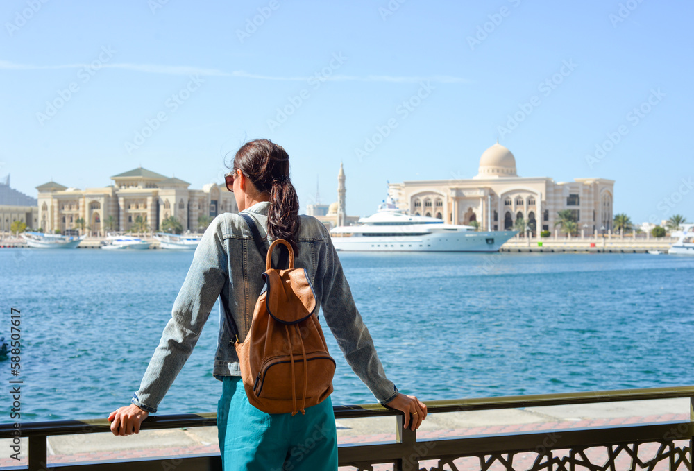 A young woman tourist with a backpack looks at the administrative region of the emirate of Sharjah with the port and ships.