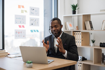 African american man in formal suit having video call at office using headset and wireless laptop. Transparent board with various graphs and charts on background. Concept of online business meeting.