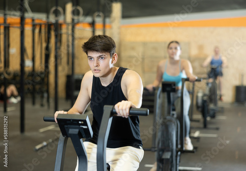 Young dedicated man training on exercise bike near other people in gym