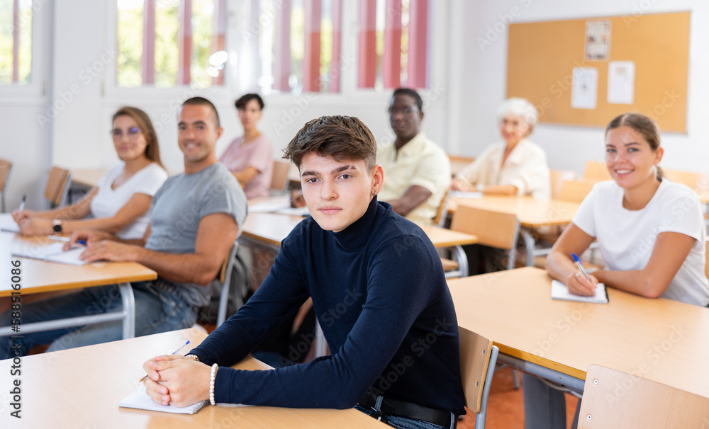Fototapeta premium Focused interested young guy attending foreign languages courses for adults, listening to lecture and taking notes while sitting at desk in auditorium