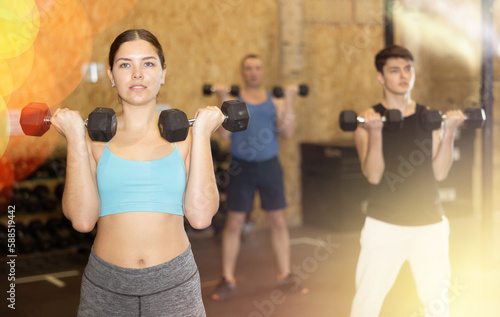 Young dedicated woman doing exercises with dumbbells near other people in gym