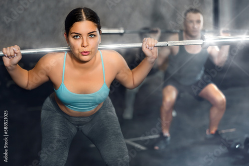 Concentrated athletic young woman taking part in intense group training in modern gym, performing exercises with barbell. Fitness and bodybuilding concept..