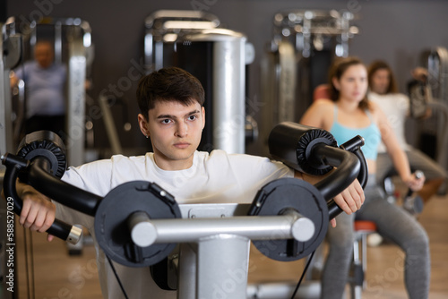 Focused young guy working out in modern gym, pumping up deltoid muscles of shoulders in lateral raise exercise machine. Active lifestyle concept and sports..