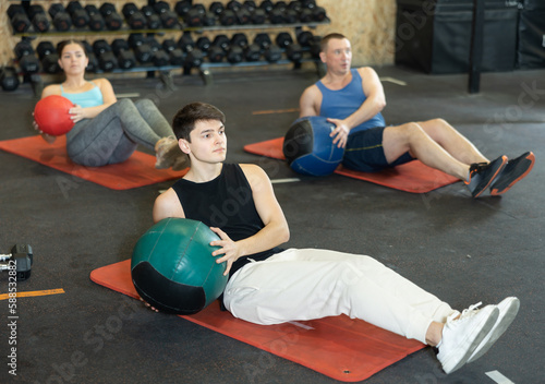 Concentrated young man in activewear working out with heavy weighted ball on mat while doing upper body training in CrossFit class