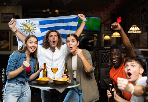 Group of Uruguay football team fans spending time in bar, drinking bear and having fun. People with state flag in pub.