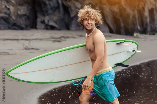 Fit young surfer man with curly blond hair with surfboard goes by the ocean having fun doing extreme water sports, surfing. Travel and healthy lifestyle concept. Sports travel destination.