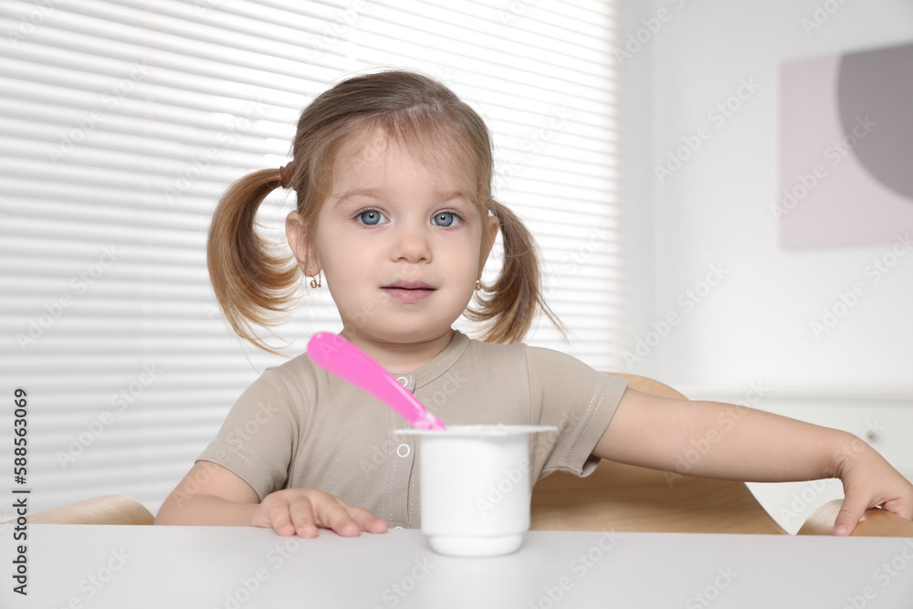 Cute little child eating tasty yogurt from plastic cup with spoon at white table indoors Cute little child eating tasty yogurt from plastic cup with spoon at white table indoors