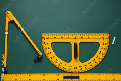 Protractor, compass, chalk and ruler on green chalkboard, flat lay