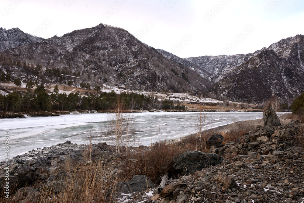 A view from a high rocky shore to the frozen bed of a beautiful river flowing at the foot of snowy mountains on a cloudy winter evening.