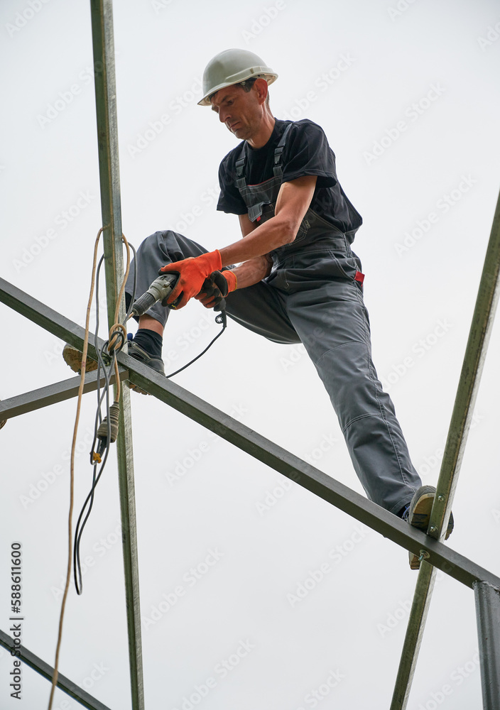 Full length of man in safety helmet and work overalls standing on metal rail and using electric drill while installing support structure for photovoltaic solar panels. Isolated on white background.