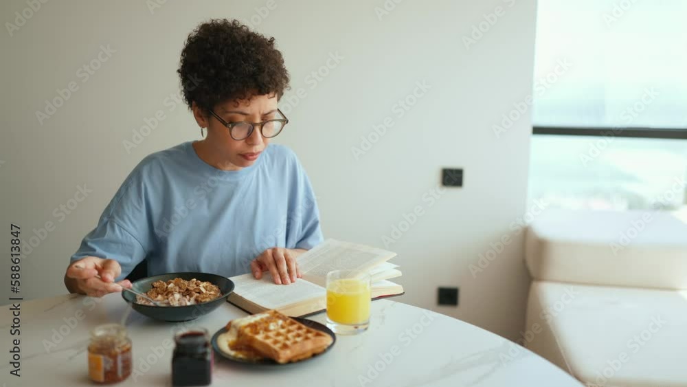 Concentrated Latin curly haired woman having breakfast and reading book in the kitchen Concentrated Latin curly haired woman having breakfast and reading book in the kitchen