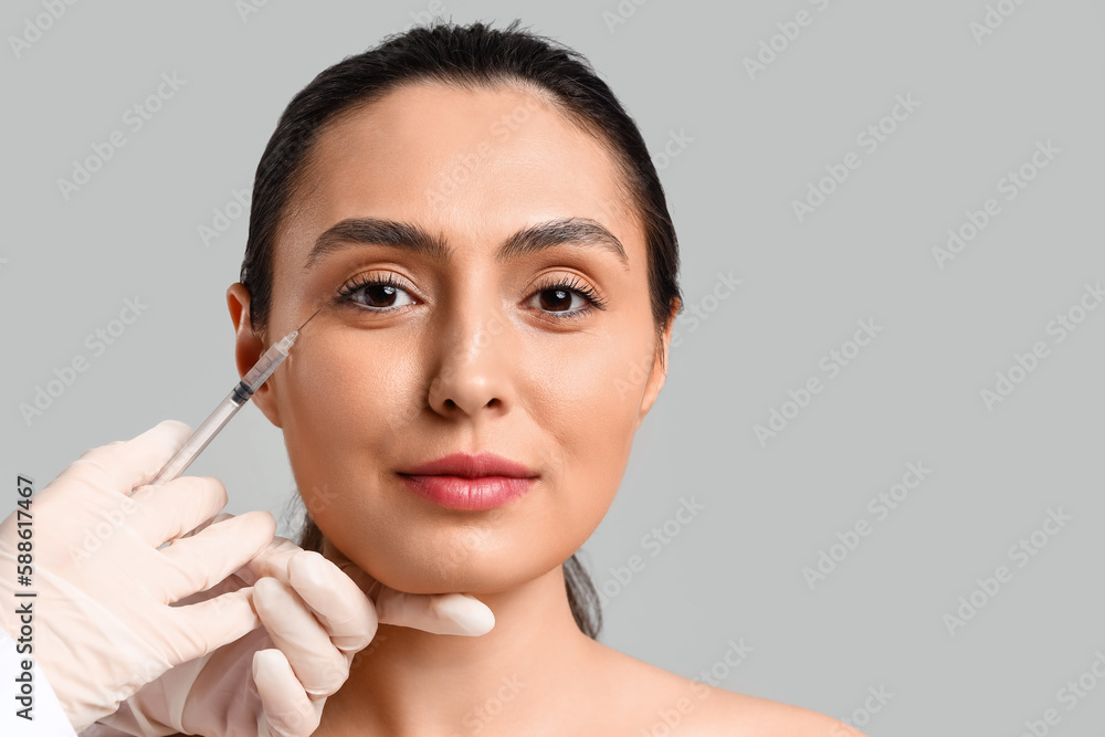 Young woman receiving filler injection in face against grey background