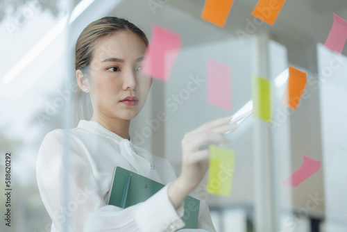 Asian woman working on project plan using sticky papers notes on glass wall, people meeting to share idea, Business design planning concepts.
