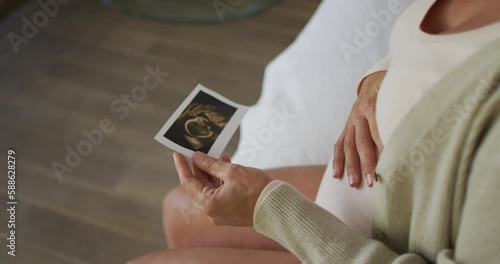 Hands of caucasian pregnant woman sitting on bed, touching belly and looking on ustrasound photo photo