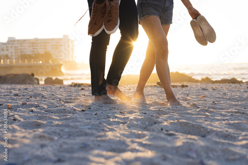 Midsection of diverse couple holding shoes and walking together at beach