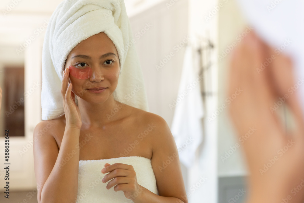 Happy asian woman wearing towel on head looking in bathroom mirror applying under eye beauty patches Happy asian woman wearing towel on head looking in bathroom mirror applying under eye beauty patches