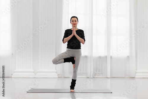 Fototapeta Naklejka Na Ścianę i Meble -  Pretty young woman doing vrikshasana on a mat on the floor in her spacious studio. Concept of benefits for the formation of correct posture heals, tones the muscles of the legs and spine