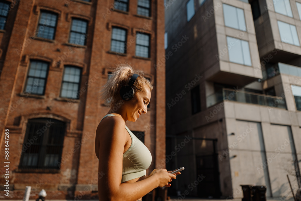 Fitness girl in sport outfit wearing headphones and turning music on smartphone. running in city