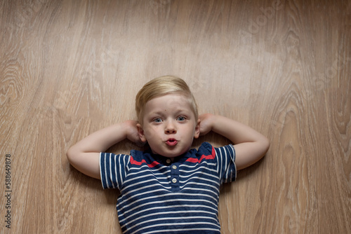 Young cute serious blond boy in stripped t-shirt is laying on the brown floor. Place for text