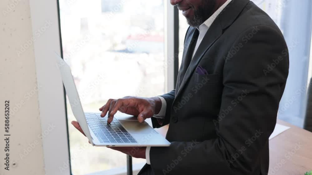 Close up of multicultural man with portable computer in hands sitting on edge of writing desk in office interior. Efficient creative manager browsing e-commerce web pages using wireless connection. Close up of multicultural man with portable computer in hands sitting on edge of writing desk in office interior. Efficient creative manager browsing e-commerce web pages using wireless connection.