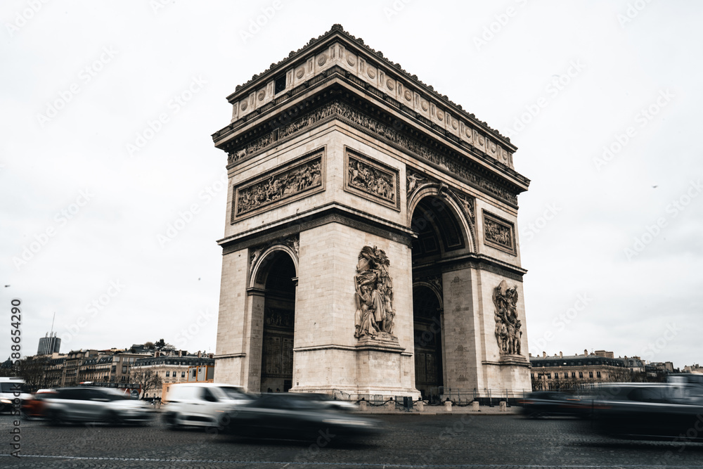 Arc de Triomphe mit Autos im Vordergrund welche in Bewegung sind und hellem bewölkten Himmel