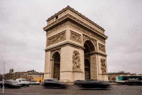 Arc de Triomphe mit Autos im Vordergrund welche in Bewegung sind und hellem bewölkten Himmel